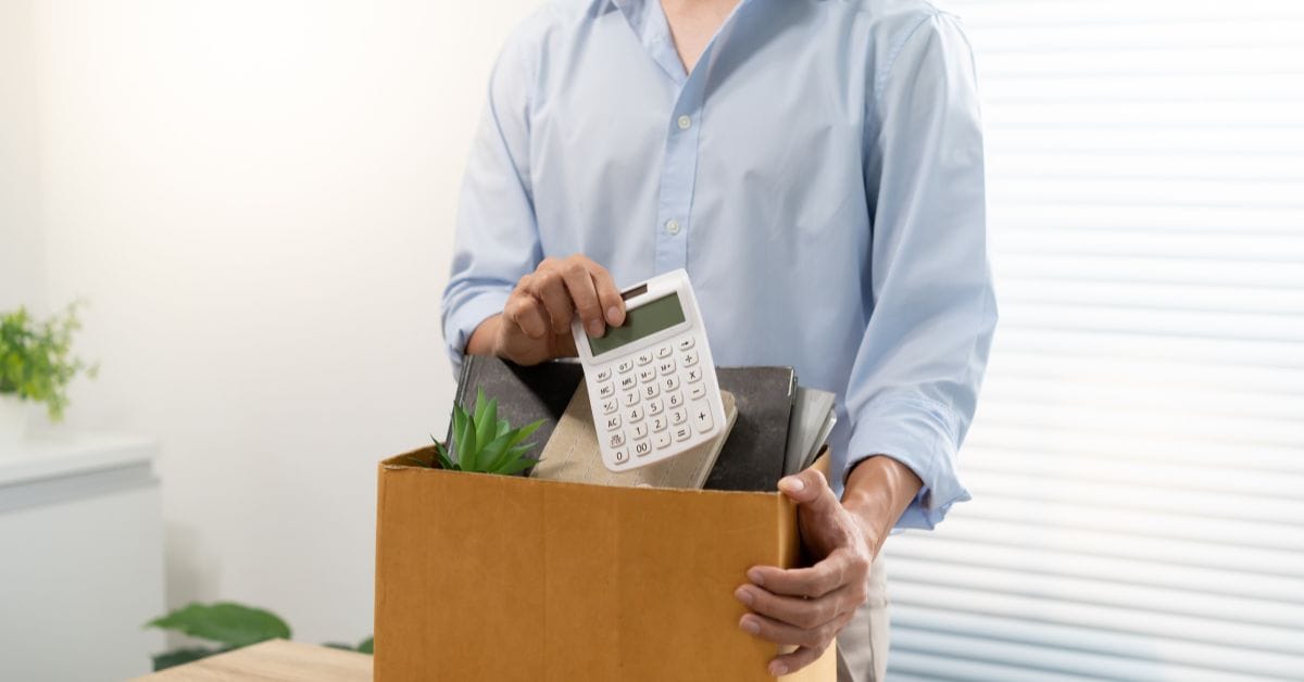 Persona empaquetando objetos personales de escritorio en una caja de cartón tras una desvinculación laboral, con una calculad